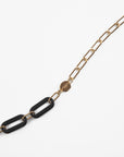 A chic, minimalist necklace featuring a chain with elongated, 18K gold-plated links. In the center are two large, black rectangular links made from Italian acetate. A small, gold circular pendant with an engraved design is attached to the chain near the black links, adding a stylish touch. This is the Kensington Glasses Chain by For Art's Sake®.