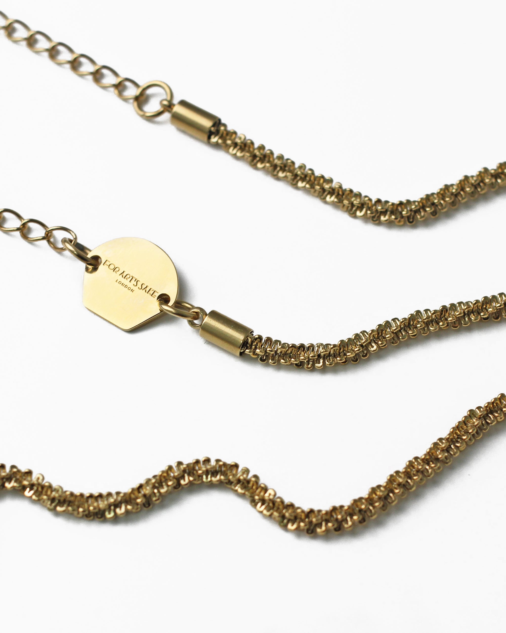 Close-up of an 18k gold-plated Mayfair Glasses Chain by For Art&#39;s Sake® featuring intricate woven links. The chain has an adjustable, heart-shaped claw closure and a small hexagonal tag engraved with the words &quot;FOR ART&#39;S SAKE.&quot; The background is white, highlighting the exquisite details of the chain and tag.