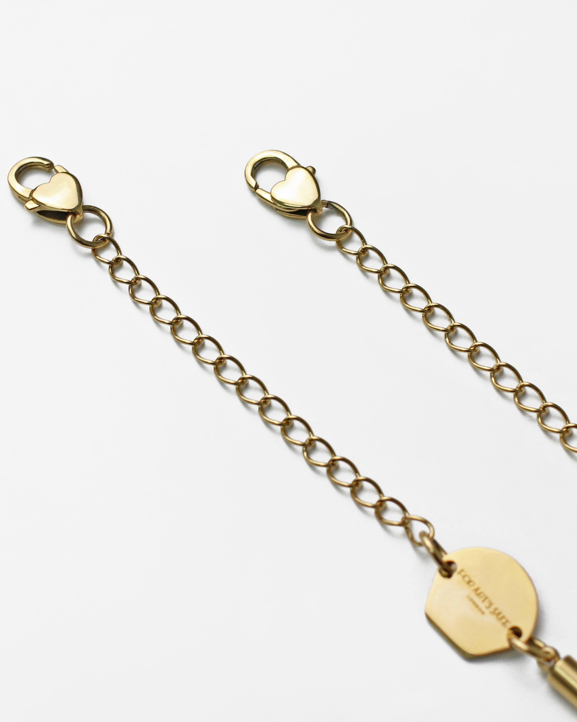 Close-up of an 18k gold-plated For Art&#39;s Sake® Mayfair Glasses Chain with circular links and two heart-shaped claw closures on each end. A small round charm bearing the word &quot;COACH&quot; is attached near one clasp. The chain lies on a plain white background.