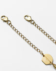 Close-up of an 18k gold-plated For Art's Sake® Mayfair Glasses Chain with circular links and two heart-shaped claw closures on each end. A small round charm bearing the word "COACH" is attached near one clasp. The chain lies on a plain white background.