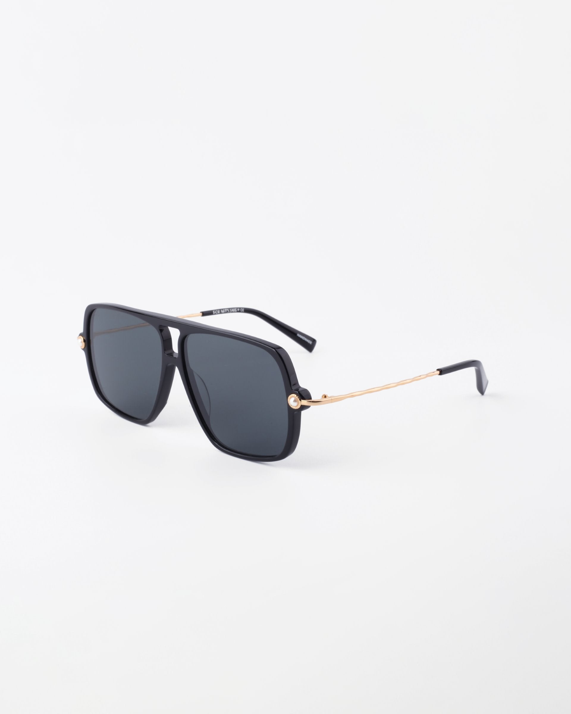 A pair of stylish Cinnamon aviator sunglasses by For Art&#39;s Sake® with black rectangular frames and dark tinted lenses. The temples are 18-karat gold-plated with a sleek design and black tips, featuring subtle faux pearl embellishment. The sunglasses are set against a white background.