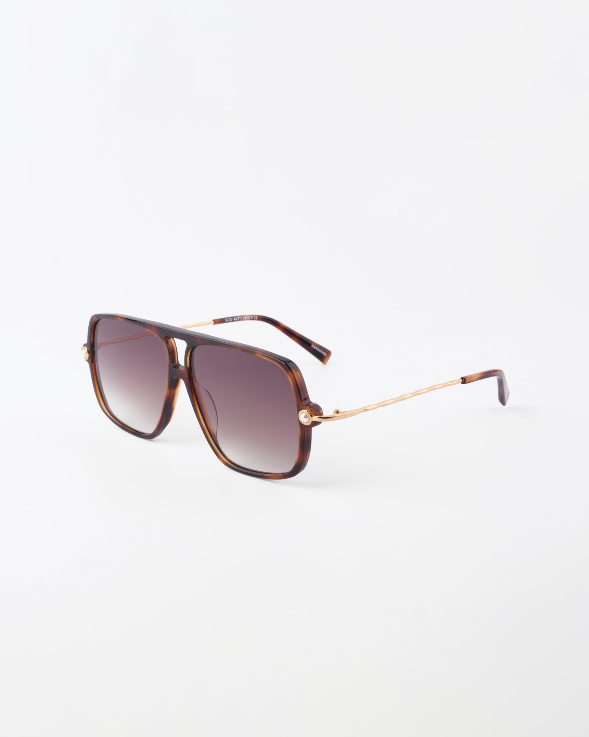 A pair of Cinnamon sunglasses by For Art&#39;s Sake® with a brown tortoiseshell frame and dark gradient lenses. The slim arms feature a gold metallic accent near the hinges, resembling 18-karat gold-plated elegance.