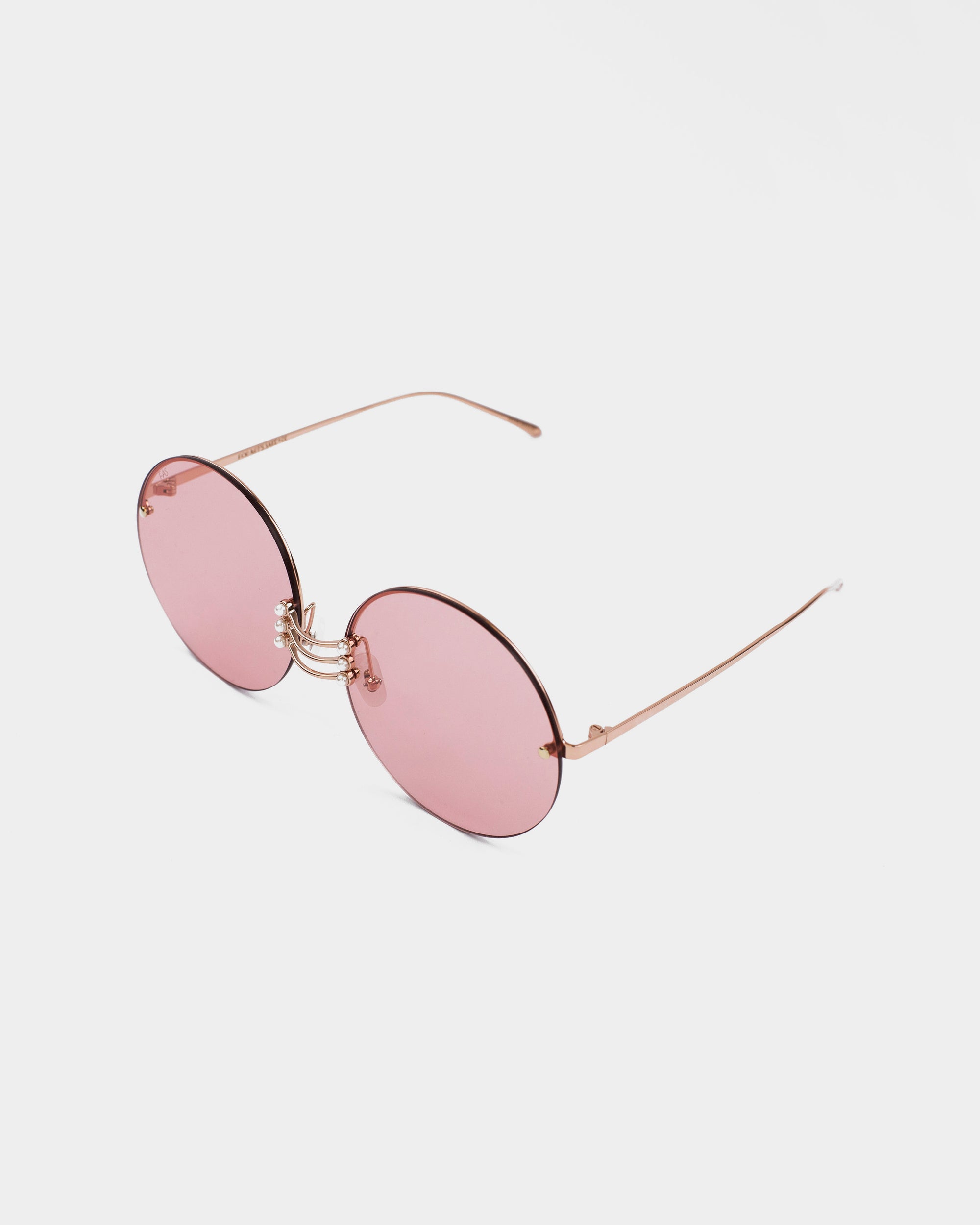 A pair of Vermeer sunglasses by For Art&#39;s Sake® with round, pink-tinted lenses and thin, gold-colored metal frames. The large lenses offer UV protection with a light pink hue. The thin arms and nose bridge are minimalist, adding an elegant touch to the eyewear&#39;s design. Stainless steel frames ensure durability, and the background is white.