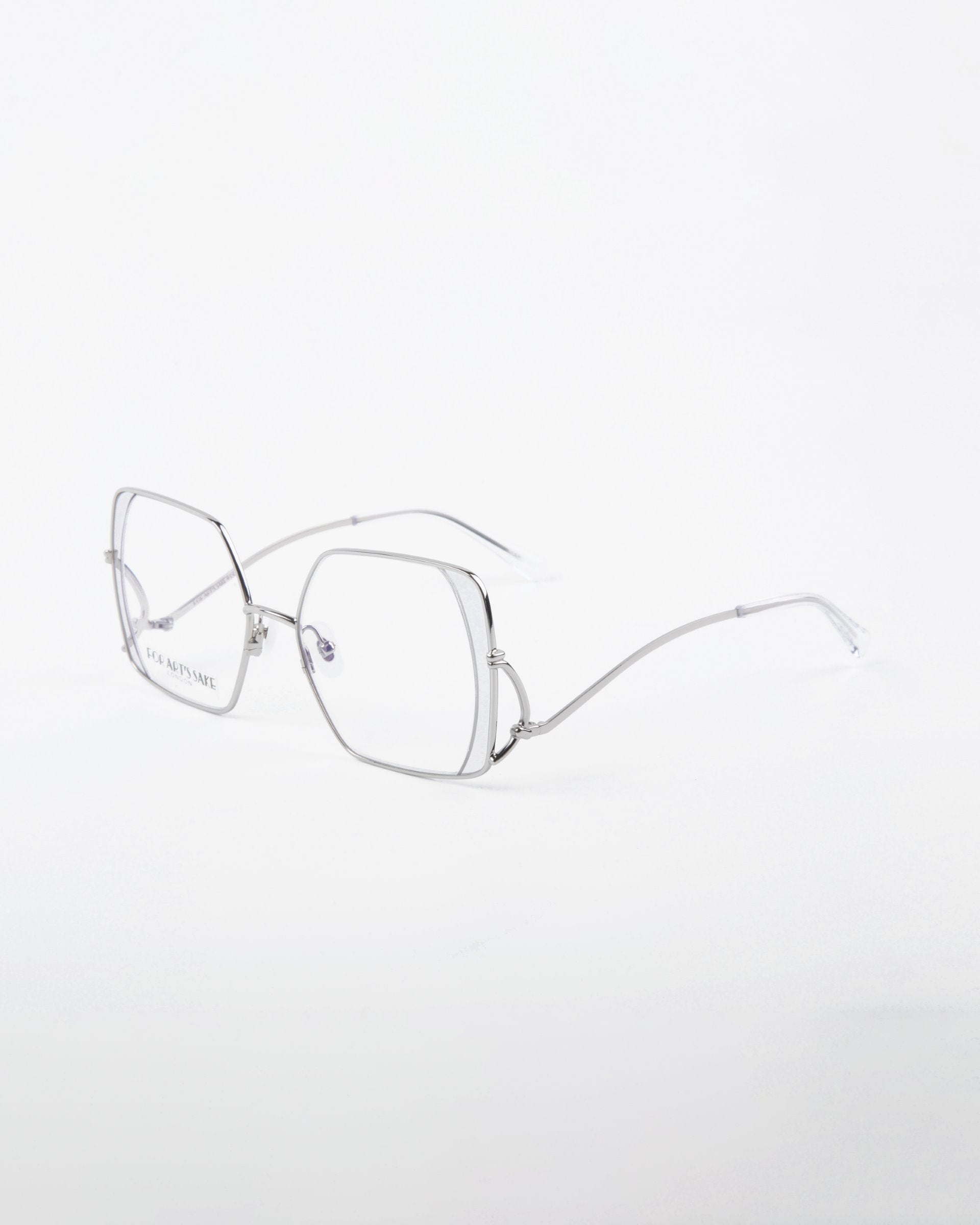 A pair of Candy eyeglasses by For Art&#39;s Sake® with silver wire frames. The lenses are slightly oversized and hexagonal, featuring a blue light filter for added eye protection. Thin stainless steel arms extend from the lenses. The design is sleek and modern, set against a plain white background.