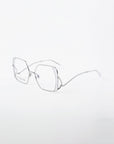 A pair of Candy eyeglasses by For Art's Sake® with silver wire frames. The lenses are slightly oversized and hexagonal, featuring a blue light filter for added eye protection. Thin stainless steel arms extend from the lenses. The design is sleek and modern, set against a plain white background.