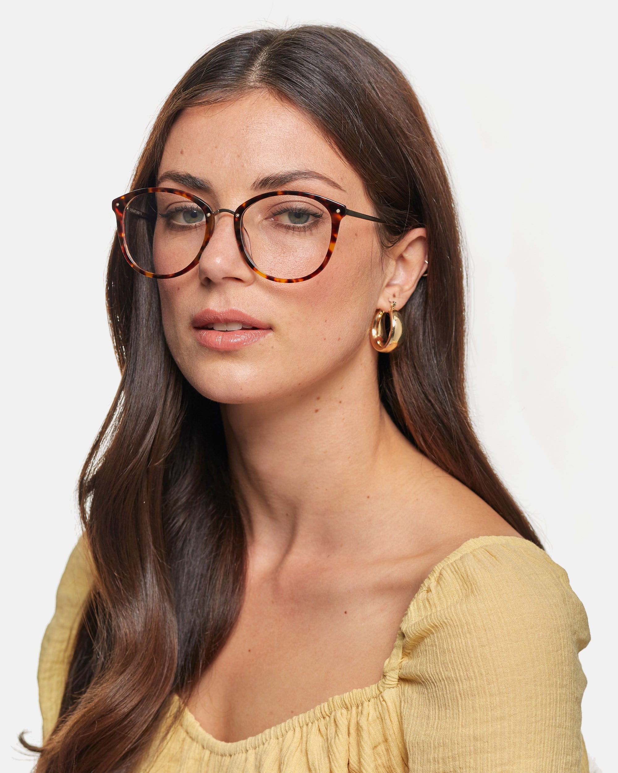 A person with long, brown hair wearing rectangular, tortoiseshell For Art&#39;s Sake® Club eyeglasses with a blue light filter and gold hoop earrings. They have on an off-the-shoulder, yellow blouse and are looking slightly to the side against a plain background.