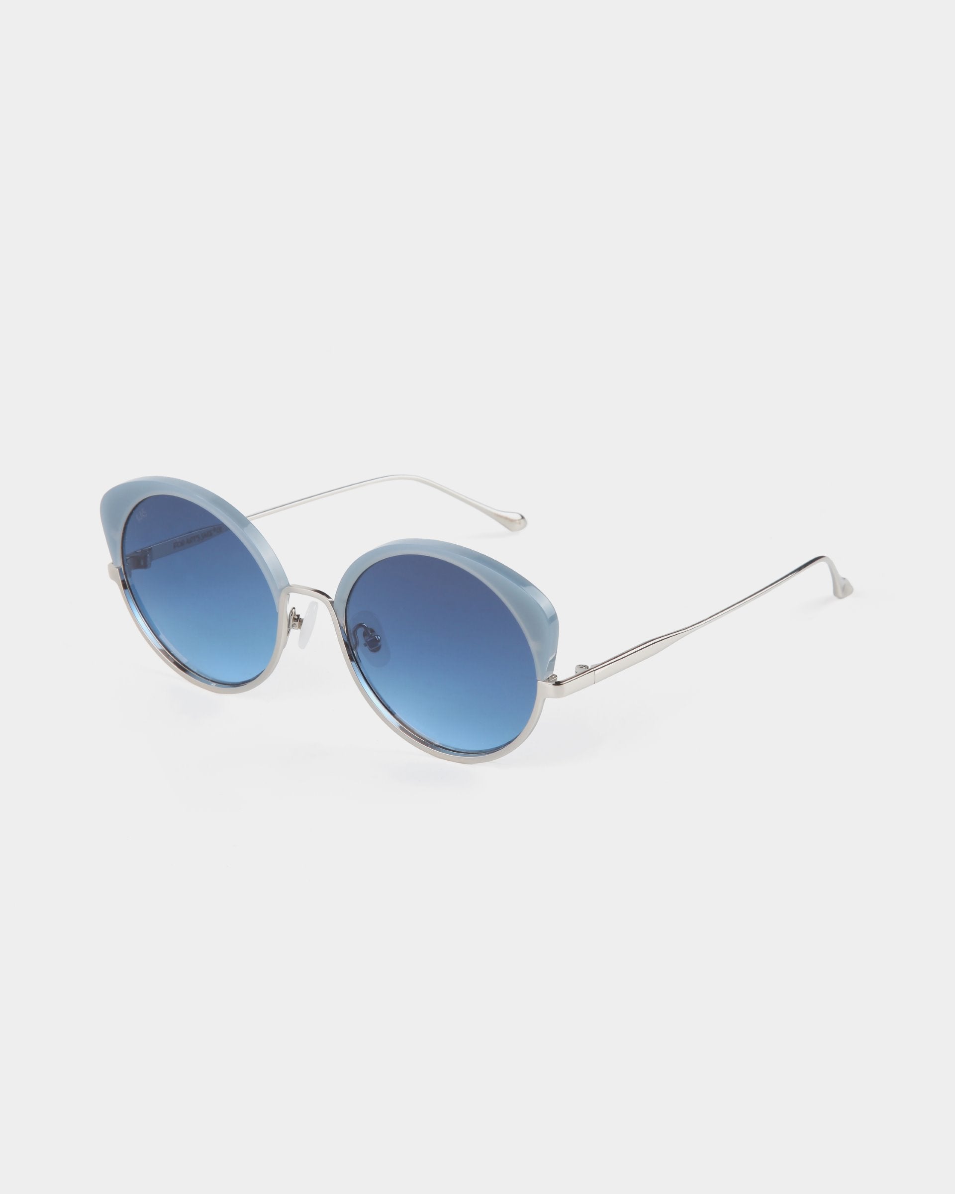 A pair of stylish Cocoon by For Art&#39;s Sake® cat-eye sunglasses with light blue, oval-shaped lenses and sleek silver frames. The arms are thin and slightly curved, featuring a minimal design. These handcrafted eyewear pieces offer UV protection and are positioned at an angle against a plain white background.