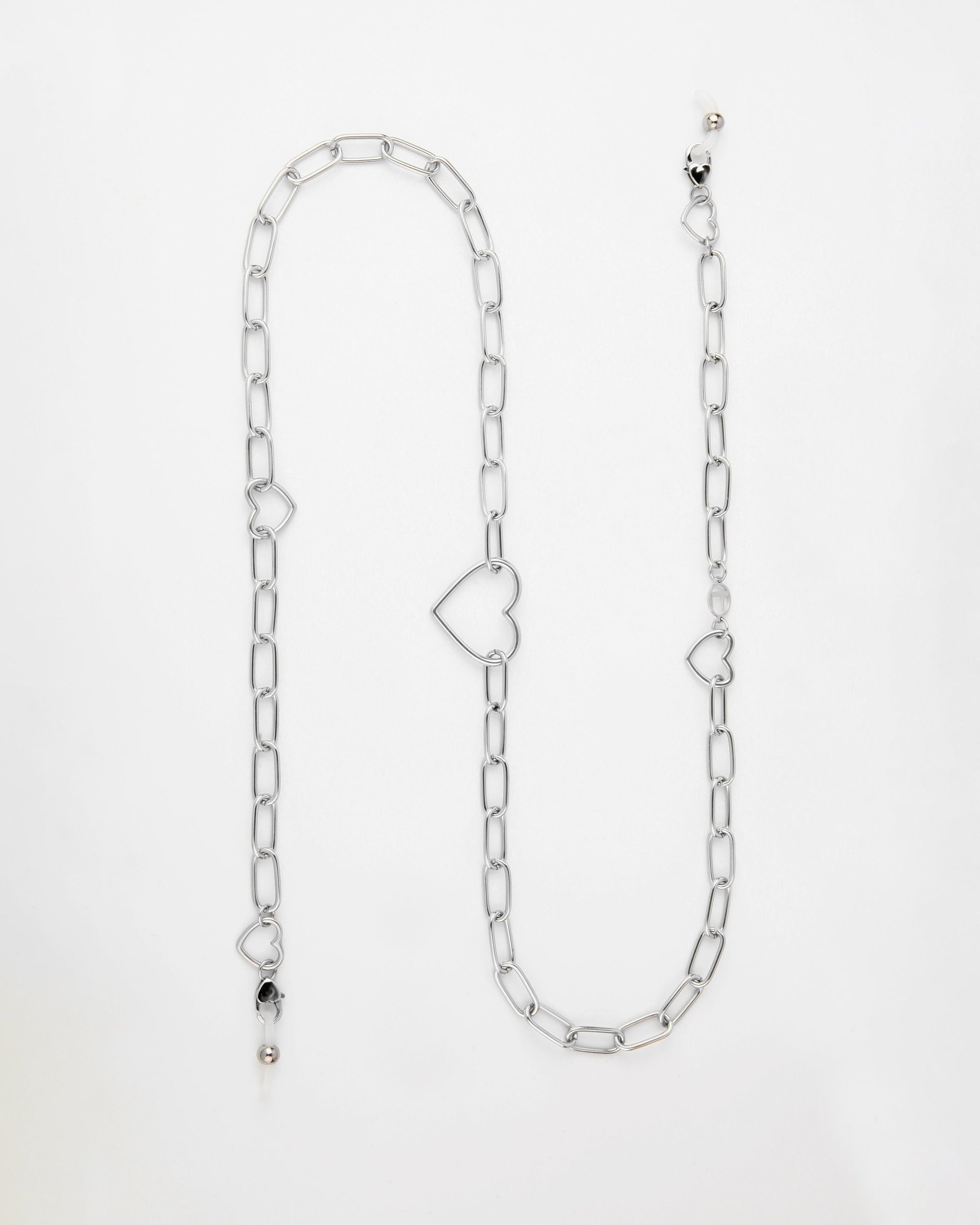 A silver Cupid Glasses Chain by For Art's Sake® with large oval links and statement hearts. The chain features a lobster clasp adorned with additional small charms, including a black bead and a pearl-like bead, attached at various points. Displayed on a plain white background, it exudes elegance and charm.