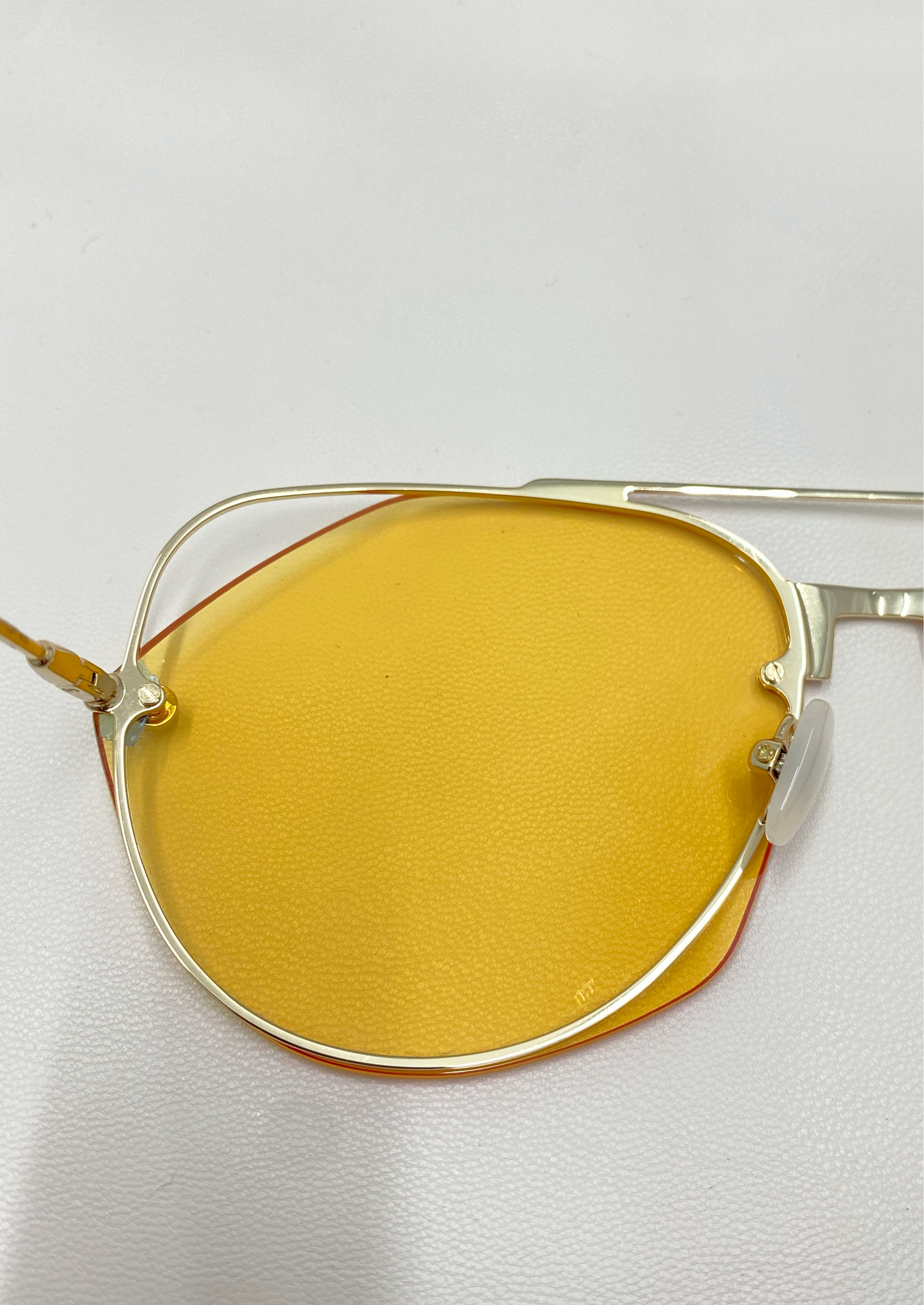 A close-up image of the Casino sunglasses by For Art&#39;s Sake® showcases modern eyewear with yellow-tinted lenses and a sleek, stainless steel frame featuring a geometric design. The background is a plain, white surface.