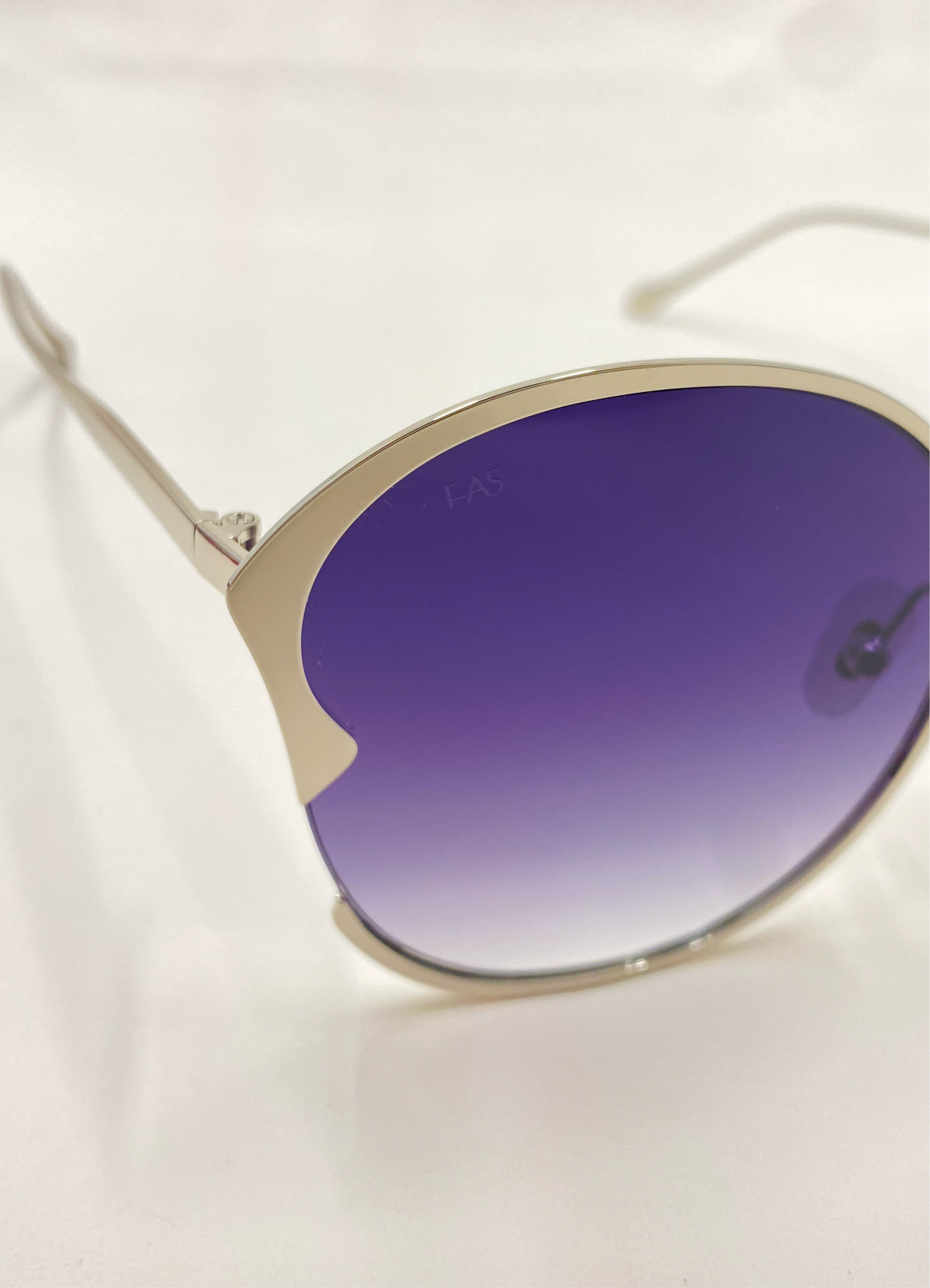 Close-up of a pair of For Art's Sake® Alectrona sunglasses featuring gradient blue, UV-protected lenses and thin, metallic gold frames. The plain white background highlights the sleek and stylish design of these sunglasses, which include a chic faux leather case.