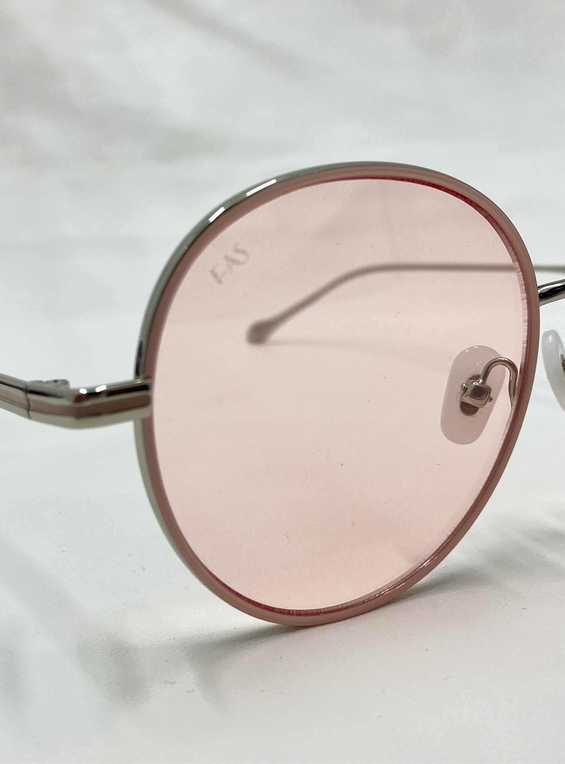 Close-up photo of For Art's Sake® Lemon sunglasses featuring round, pink-tinted lenses in thin metallic frames with clear jadestone nose pads. The slightly reflective lenses display a subtle "FAS" marking on the left lens. The background is a plain, light-colored surface.