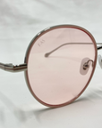 Close-up photo of For Art's Sake® Lemon sunglasses featuring round, pink-tinted lenses in thin metallic frames with clear jadestone nose pads. The slightly reflective lenses display a subtle "FAS" marking on the left lens. The background is a plain, light-colored surface.