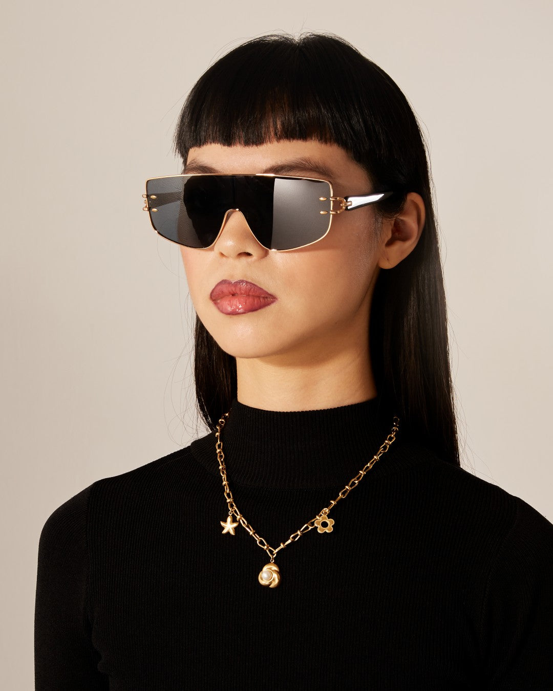 A woman with long black hair and bangs is wearing oversized black sunglasses and a black turtleneck. She sports the Daisy Necklace Gold, a gold plated stainless steel chain necklace with star and sun charms, from For Art&#39;s Sake®. The background is off-white, and her expression is neutral.