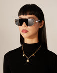 A woman with long black hair and bangs is wearing oversized black sunglasses and a black turtleneck. She sports the Daisy Necklace Gold, a gold plated stainless steel chain necklace with star and sun charms, from For Art's Sake®. The background is off-white, and her expression is neutral.