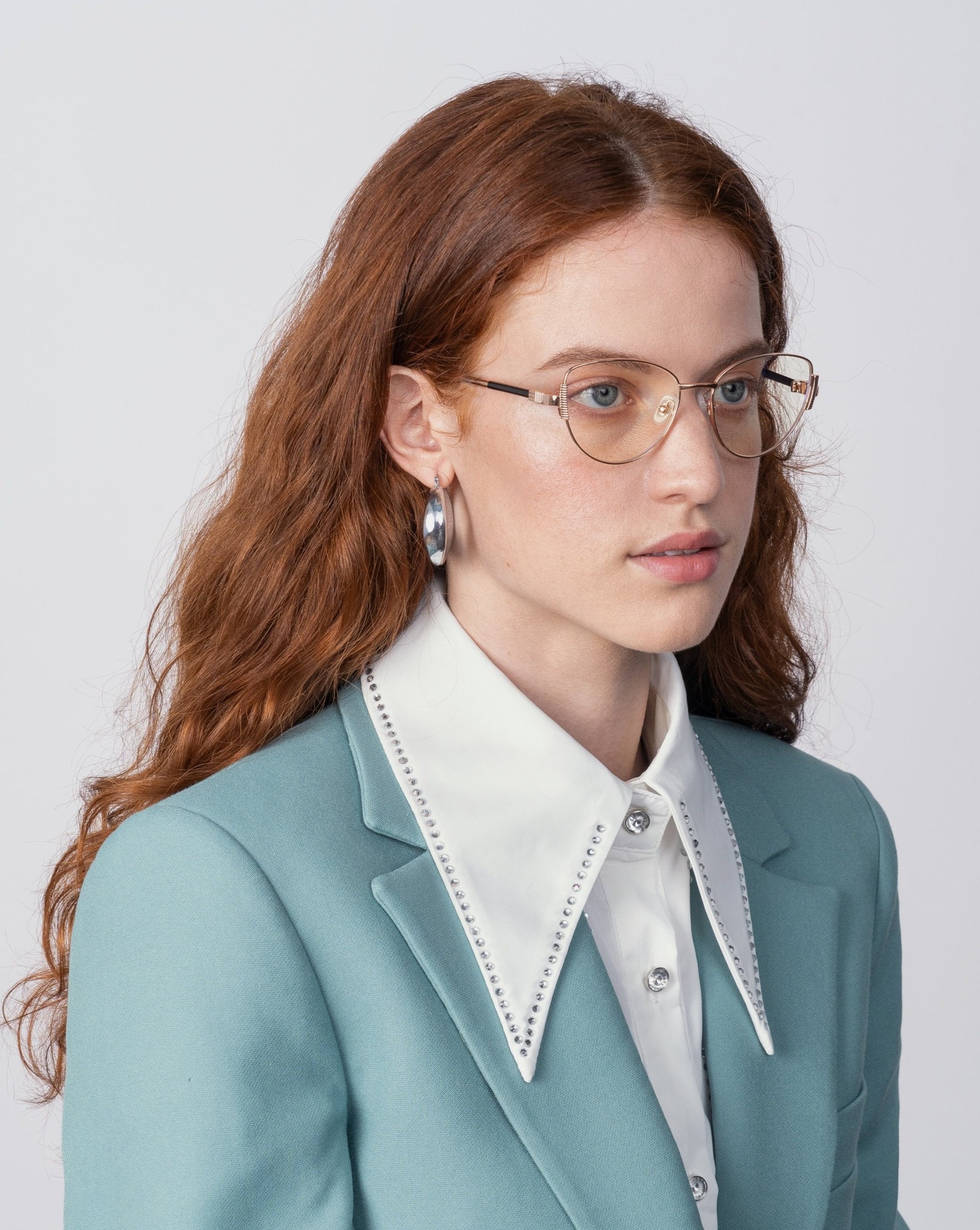 A woman with long, wavy red hair is wearing Dante eyeglasses from For Art's Sake® with blue light filter lenses, silver hoop earrings, and a light blue blazer over a white shirt with a wide, pointed collar. She is looking slightly to the side against a plain, light background.