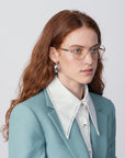 A woman with long, wavy red hair is wearing Dante eyeglasses from For Art's Sake® with blue light filter lenses, silver hoop earrings, and a light blue blazer over a white shirt with a wide, pointed collar. She is looking slightly to the side against a plain, light background.