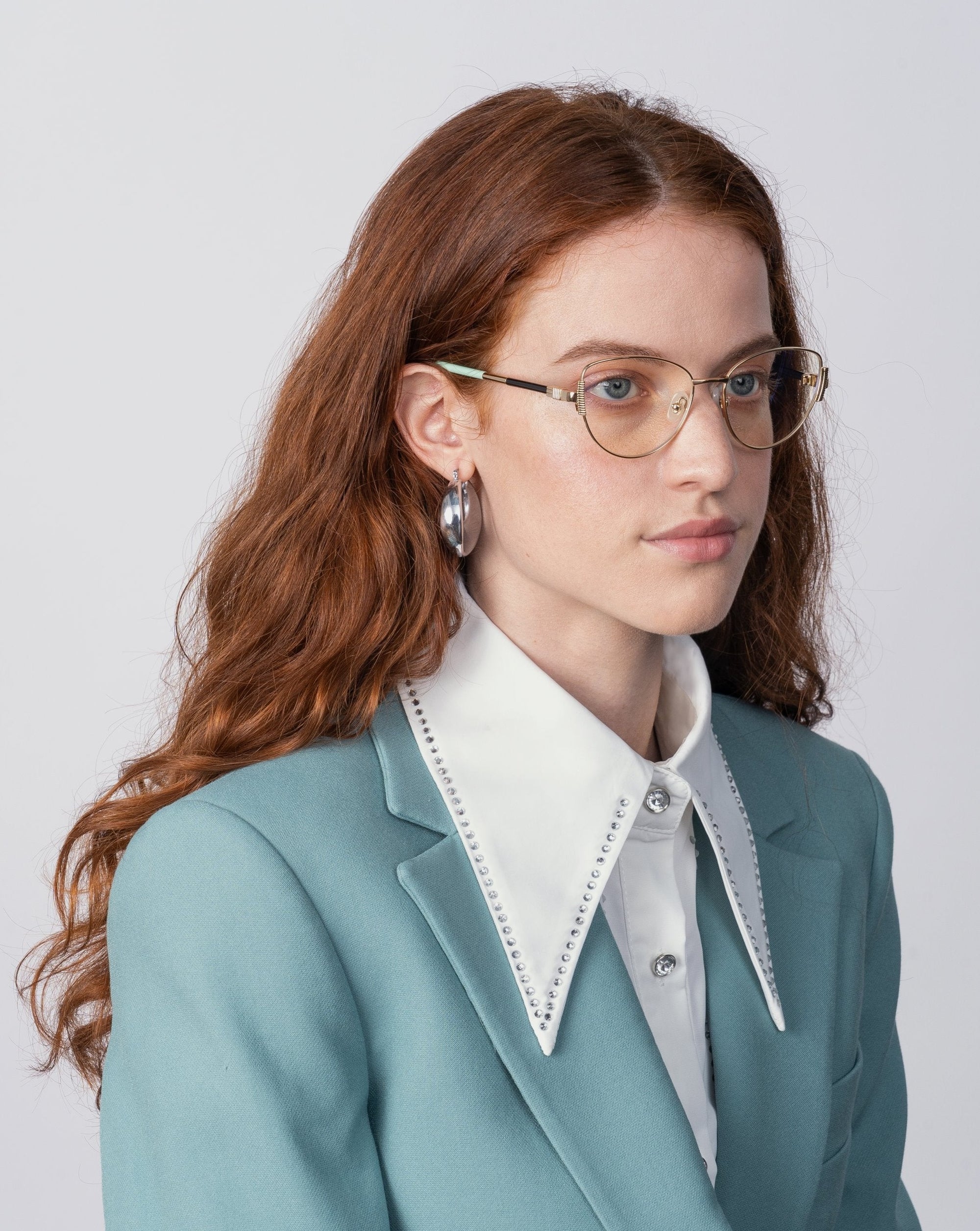 A person with long, wavy auburn hair wears a teal blazer over a white shirt with a large collar and metallic studs. They have glasses with prescription lenses and jade stone nose pads from the brand For Art's Sake®, specifically the Dante model, along with silver hoop earrings. They're looking to the side against a plain white background.