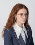 A person with long, wavy red hair, wearing Dante by For Art's Sake® with jade stone nose pads and a blue light filter, pearl earrings, and a dark blazer with white trim over a white shirt with an exaggerated collar, poses against a plain background.