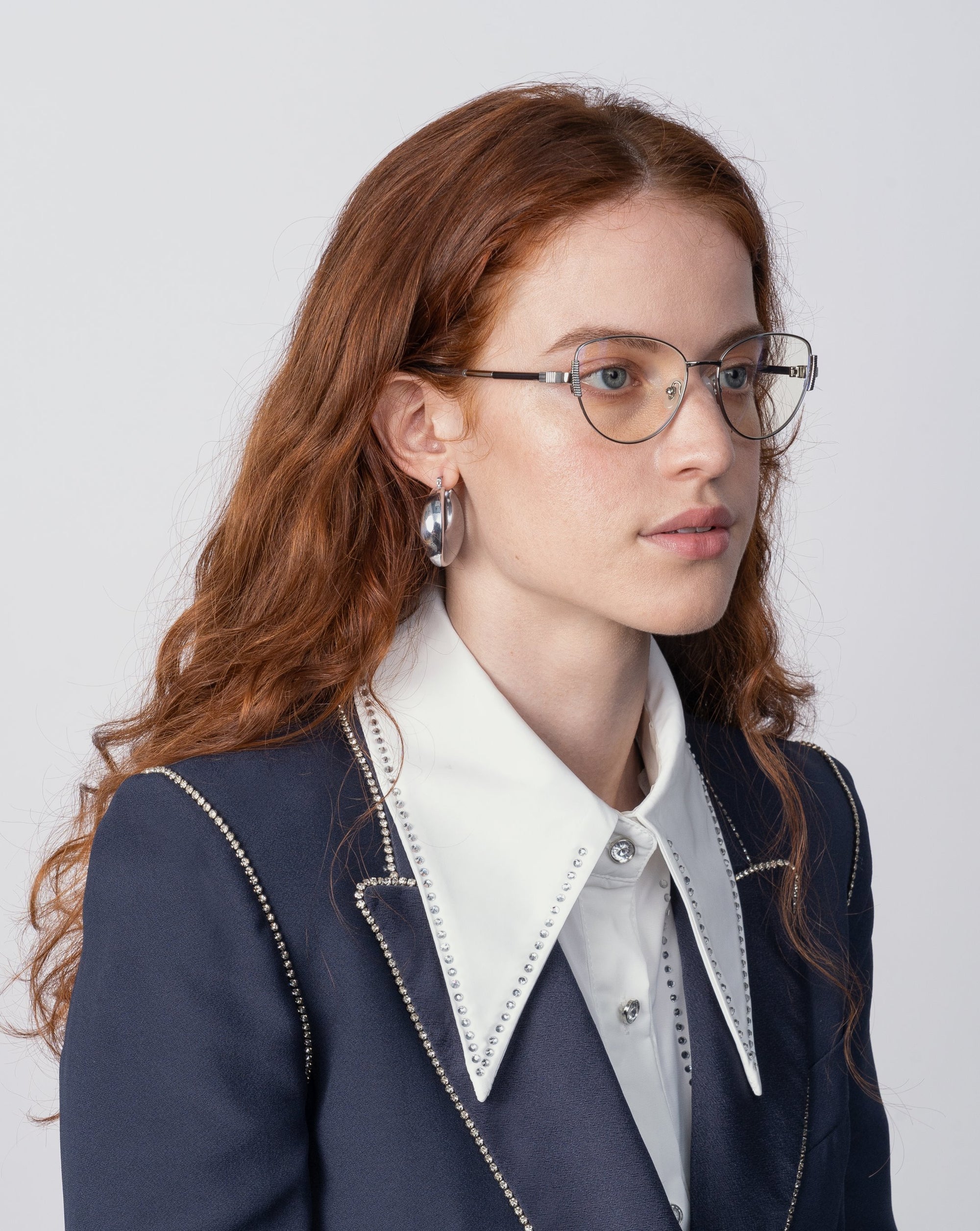 A person with long red hair and glasses with For Art&#39;s Sake® Dante jade stone nose pads is pictured against a plain background. They are wearing a dark blazer with white studs, a white shirt featuring a large collar, and silver hoop earrings.