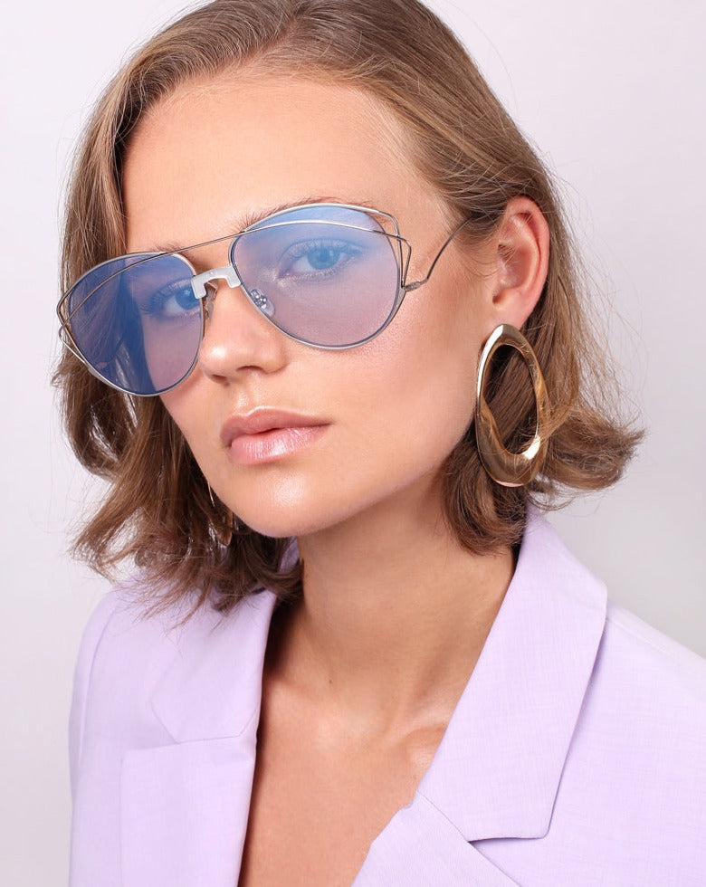 A person with short, wavy hair is wearing large, blue-tinted Dark Eyes sunglasses by For Art's Sake® with 100% UV protection and oversized, gold hoop earrings. They are dressed in a light lavender blazer and posing against a plain background.