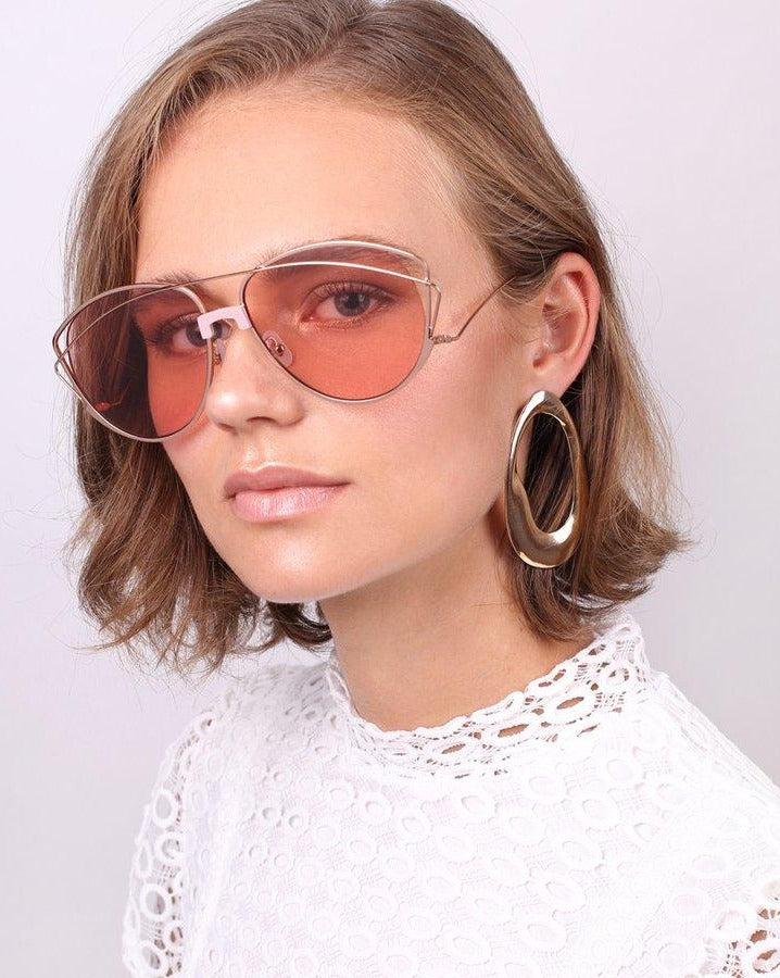 A woman with short, light brown hair is wearing large, pink-tinted Dark Eyes aviator sunglasses with nylon lenses and 100% UV protection from For Art&#39;s Sake®, paired with a white lace top. She has on large hoop earrings and is gazing confidently at the camera against a plain, light background.