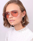 A woman with short, light brown hair is wearing large, pink-tinted Dark Eyes aviator sunglasses with nylon lenses and 100% UV protection from For Art's Sake®, paired with a white lace top. She has on large hoop earrings and is gazing confidently at the camera against a plain, light background.