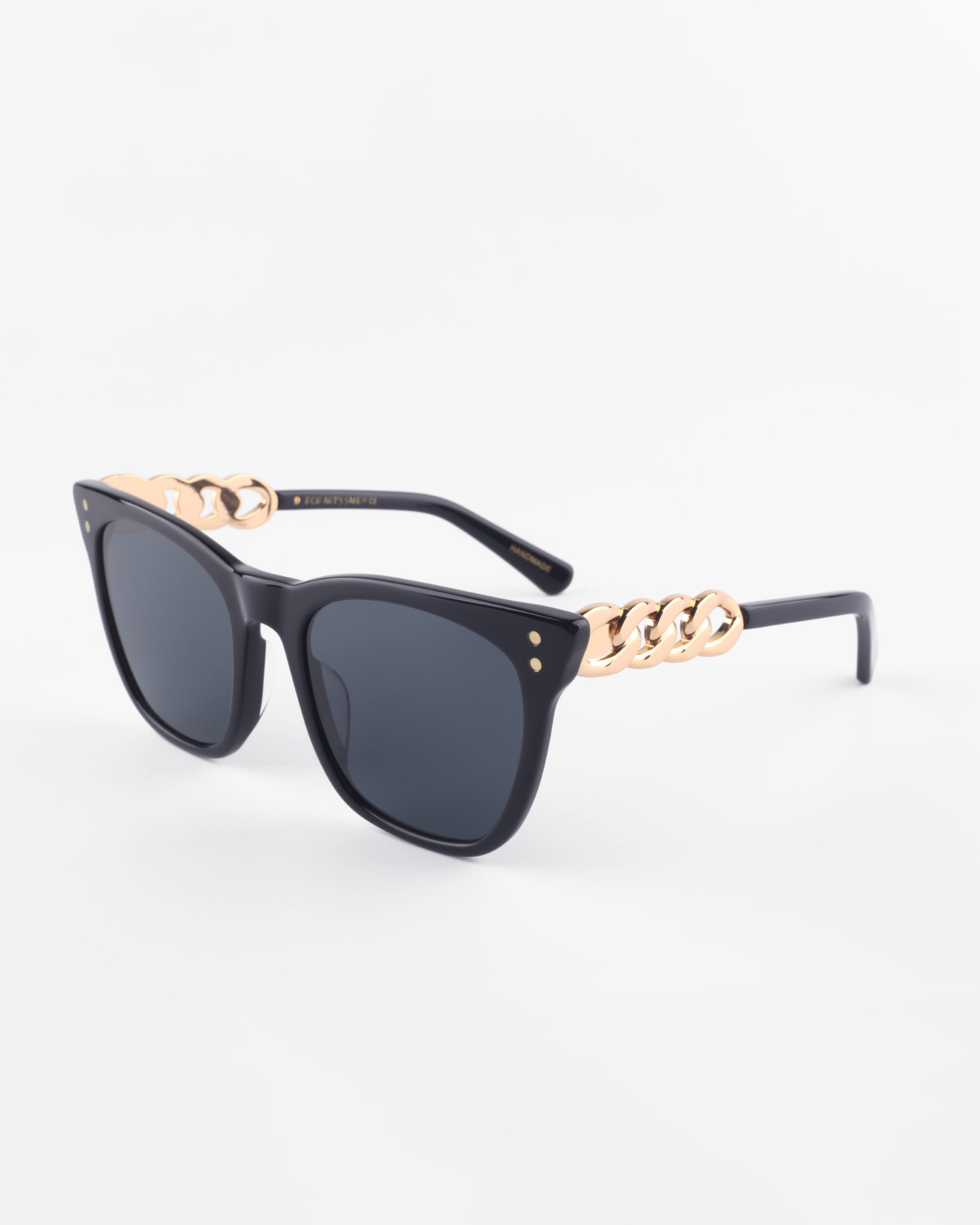 A pair of stylish black cat-eye Deco sunglasses with dark lenses from For Art's Sake®. The acetate temples feature a unique design with chunky gold chain links near the hinges, adding a touch of luxury to the classic black frames. The background is plain white.