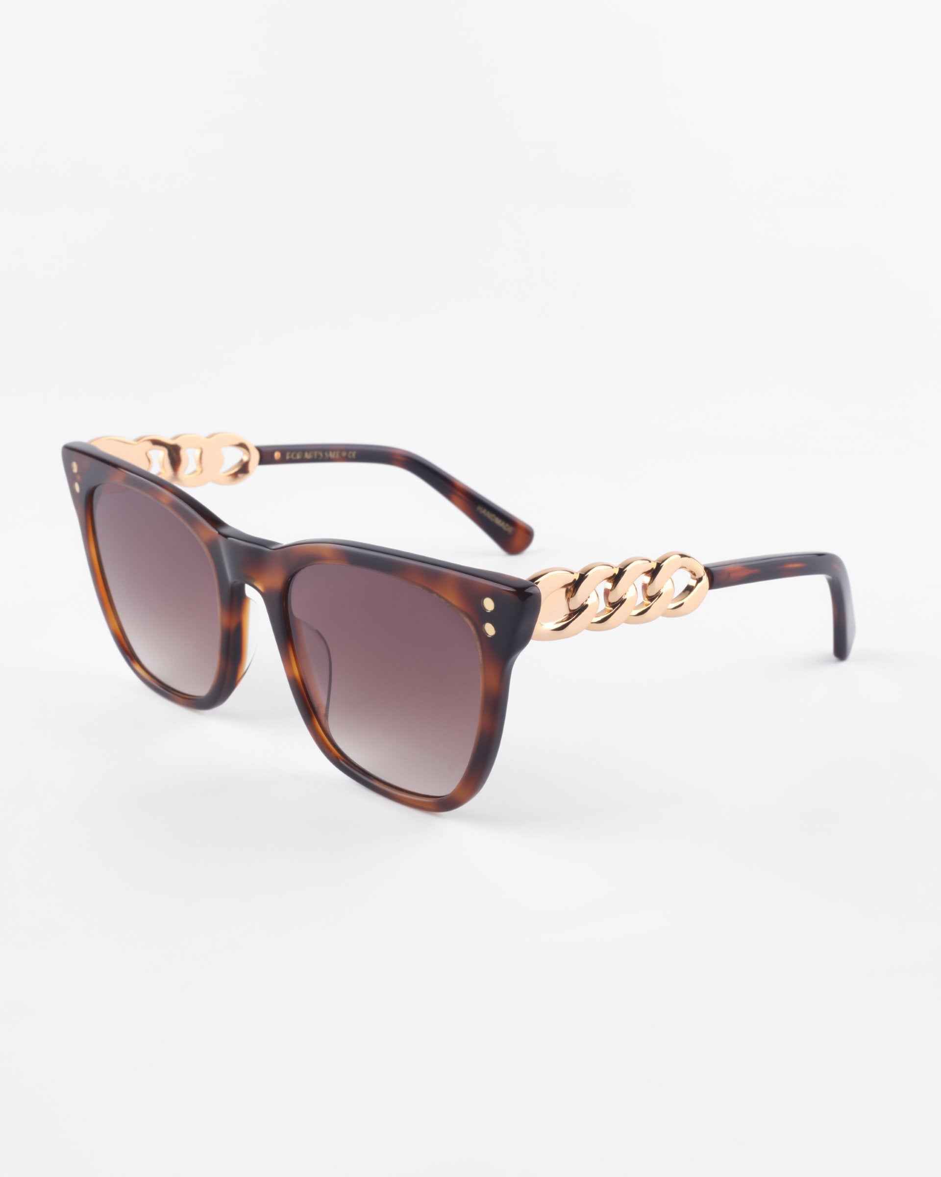 A pair of stylish Deco cat-eye sunglasses by For Art&#39;s Sake® with tortoiseshell acetate frames and dark lenses. The arms of the Deco sunglasses feature a distinctive gold chain link design near the hinges. The background is plain white.