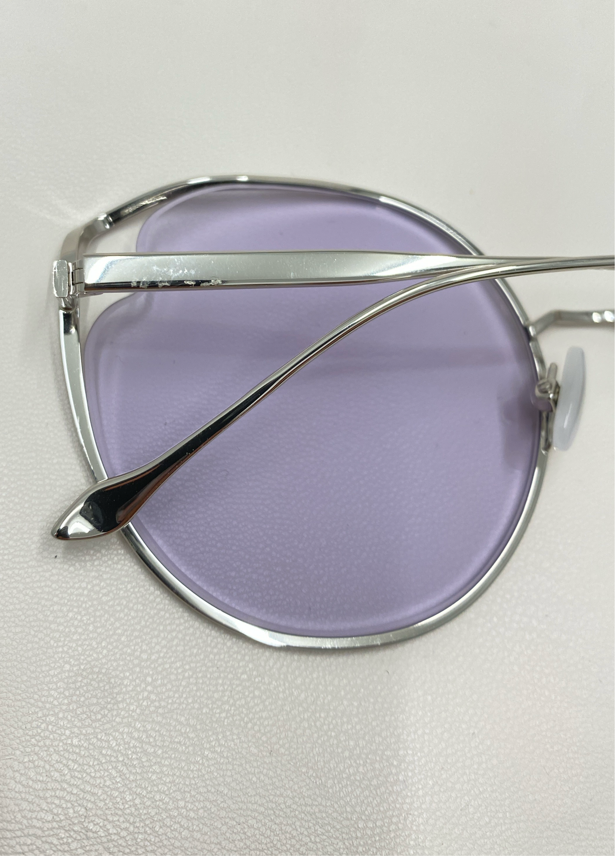 Close-up of a pair of For Art&#39;s Sake® Cat sunglasses featuring light purple tinted nylon lenses and silver metallic frames. The image focuses on the side profile, highlighting the sleek design, UVA &amp; UVB-protected lenses, and thin earpieces resting on a smooth, white surface.