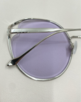 Close-up of a pair of For Art's Sake® Cat sunglasses featuring light purple tinted nylon lenses and silver metallic frames. The image focuses on the side profile, highlighting the sleek design, UVA & UVB-protected lenses, and thin earpieces resting on a smooth, white surface.