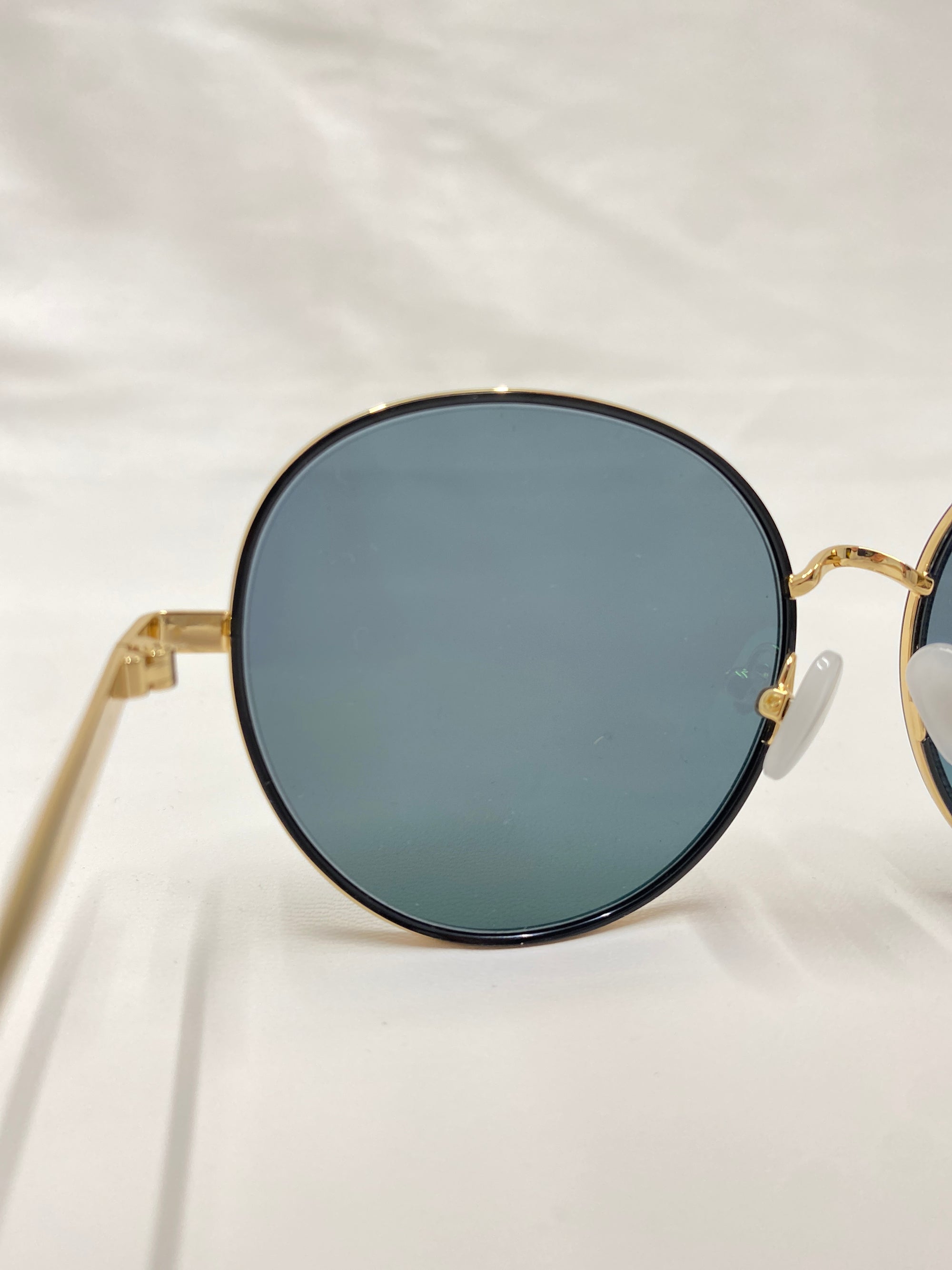 Close-up of a pair of Lemon sunglasses by For Art&#39;s Sake®, featuring round, gold-framed design with dark tinted lenses and jadestone nose pads. The background is a plain white surface, highlighting one lens and part of the frame prominently.