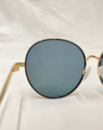 Close-up of a pair of Lemon sunglasses by For Art's Sake®, featuring round, gold-framed design with dark tinted lenses and jadestone nose pads. The background is a plain white surface, highlighting one lens and part of the frame prominently.