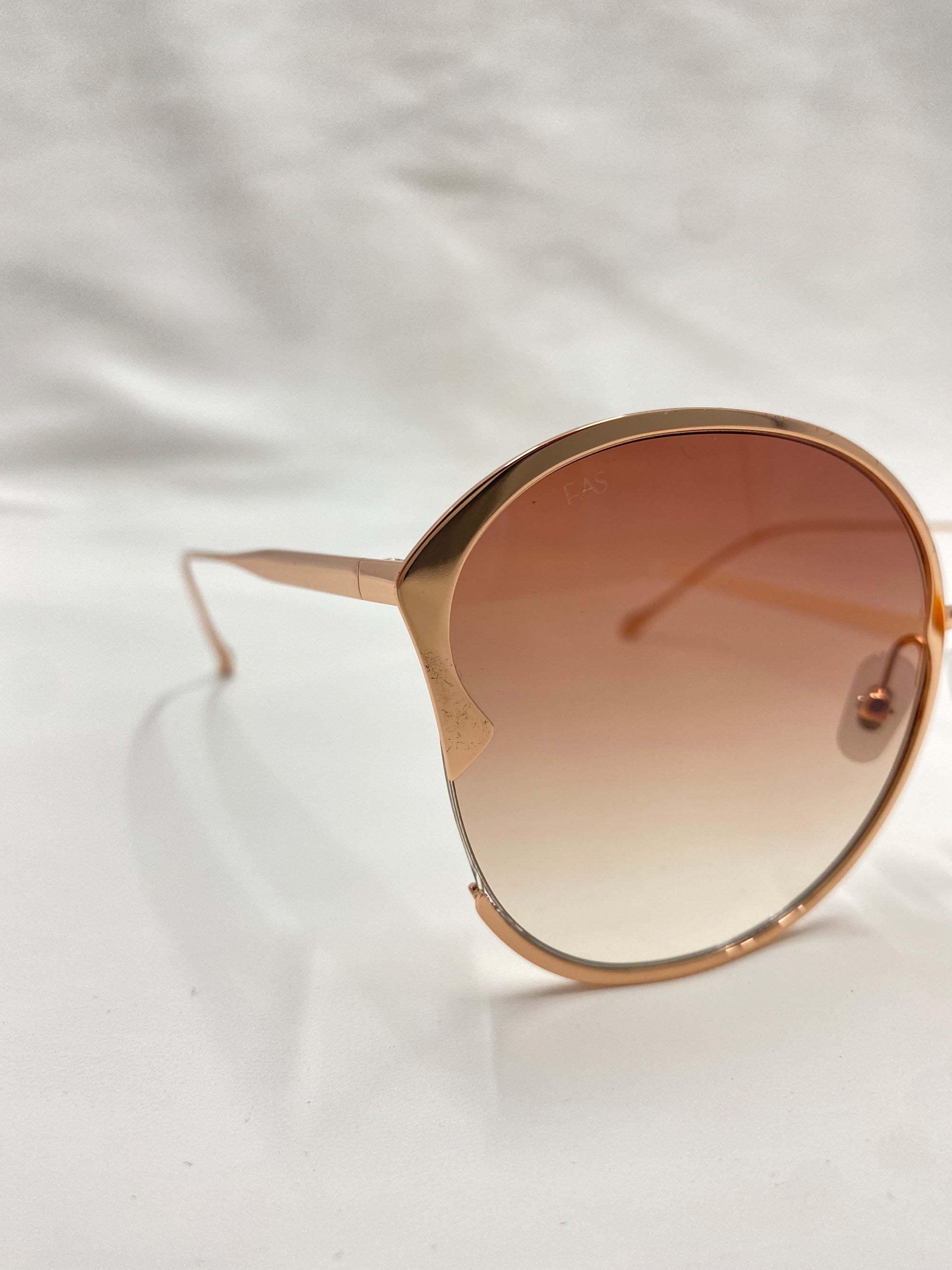 A close-up image of For Art&#39;s Sake® Alectrona sunglasses featuring large, round gradient lenses and thin, metallic frames. The UVA &amp; UVB-protected lenses ensure maximum eye safety. The background is an off-white or light cream color, with the focus on the left side of the Alectrona sunglasses.