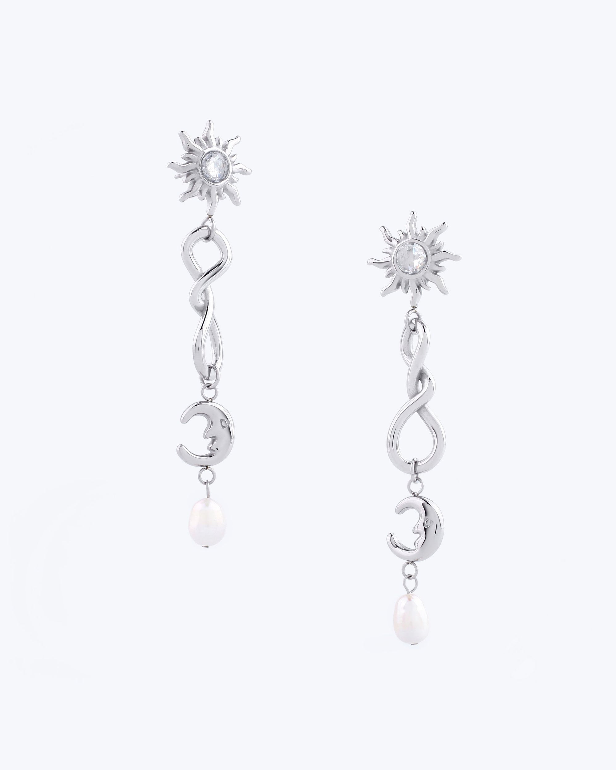 The Sloane Earrings by For Art&#39;s Sake® are exquisite silver pieces featuring a sun-shaped stud adorned with a central gemstone. Each earring showcases a wavy design that flows into a crescent moon charm, culminating in a small dangling pearl. Displayed on a light background, these earrings perfectly blend elegance and charm.