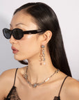 A person with long dark hair wears black sunglasses, a black spaghetti-strap top, and a silver chain necklace adorned with pearls. Their standout accessory is the For Art's Sake® Sloane Earrings, featuring stars and pearls. The plain, light-colored background enhances the ensemble's elegance.