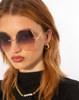 A person with long, light brown hair is wearing oversized, gold-plated stainless steel For Art's Sake® Generation sunglasses with tinted, ultra-lightweight nylon lenses that offer UV protection. They have on hoop earrings and a pearl necklace, dressed in a black top and facing the camera against a white background.