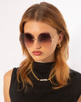 A young woman with medium-length reddish-brown hair wears large, hexagonal sunglasses called Generation by For Art's Sake® with UV protection and gradient lenses in gold-plated stainless steel frames. She has a serious expression and is dressed in a black sleeveless top, accessorized with gold hoop earrings and a gold chain necklace with a pearl.