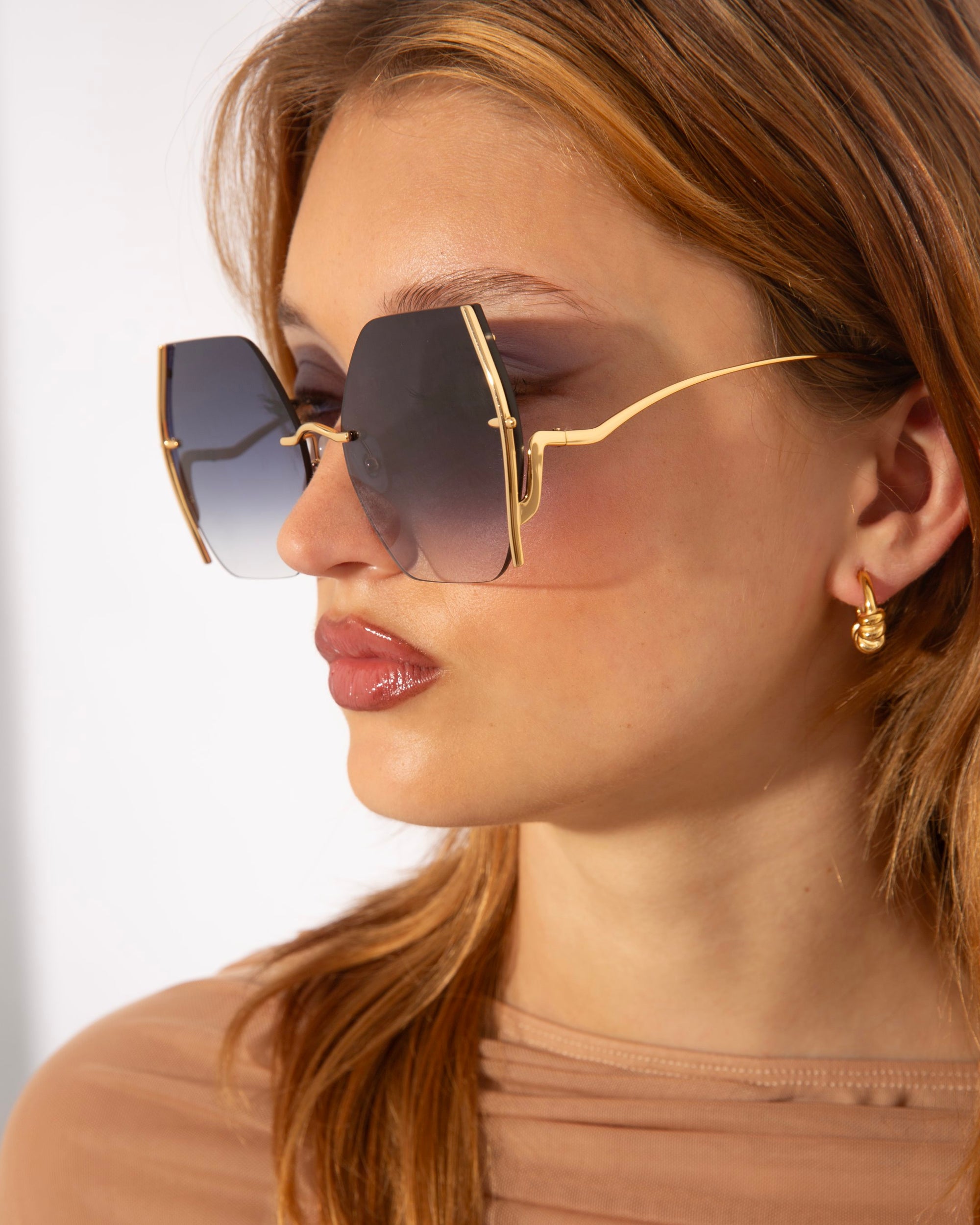 A person with long, light brown hair wears large, geometric-shaped gradient For Art&#39;s Sake® Generation sunglasses featuring jadestone nosepads and small, gold earrings. They have a neutral expression and are dressed in a light brown, semi-sheer top, shown in a side profile against a plain background.