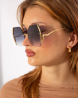 A person with long, light brown hair wears large, geometric-shaped gradient For Art's Sake® Generation sunglasses featuring jadestone nosepads and small, gold earrings. They have a neutral expression and are dressed in a light brown, semi-sheer top, shown in a side profile against a plain background.