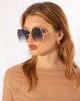 A woman with wavy, reddish-brown hair is wearing oversized Generation sunglasses by For Art's Sake® with a thin, gold-plated stainless steel frame and jadestone nosepads. She has on gold hoop earrings and a sheer, beige top. The background is plain white.