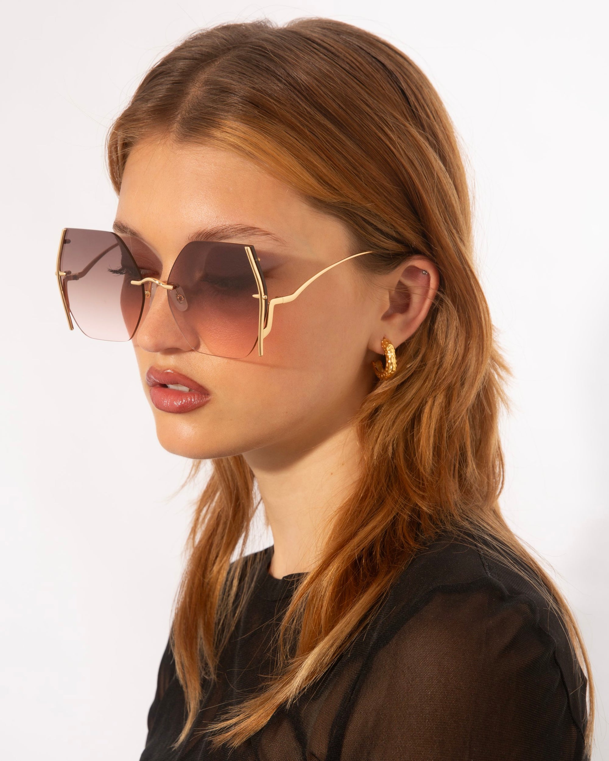 A person with long, light brown hair is wearing large, square-shaped For Art&#39;s Sake® Generation sunglasses with gold accents and gradient lenses offering UV protection. They are also wearing a black, semi-transparent top and a gold hoop earring. The background is plain white.