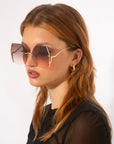 A person with long, light brown hair is wearing large, square-shaped For Art's Sake® Generation sunglasses with gold accents and gradient lenses offering UV protection. They are also wearing a black, semi-transparent top and a gold hoop earring. The background is plain white.