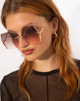 A person with long, light brown hair wears oversized, square-shaped sunglasses with gradient-tinted lenses that offer UV protection. They are also wearing gold hoop earrings and a sheer black top, looking slightly downwards against a neutral background. The For Art's Sake® Generation sunglasses feature elegant gold-plated stainless steel frames.