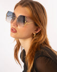 A person with long, wavy auburn hair wears large, fashionable For Art's Sake® Generation sunglasses with gold frames and gradient lenses equipped with UV protection. They have light skin, are wearing a black top, and have gold hoop earrings. The background is plain white.