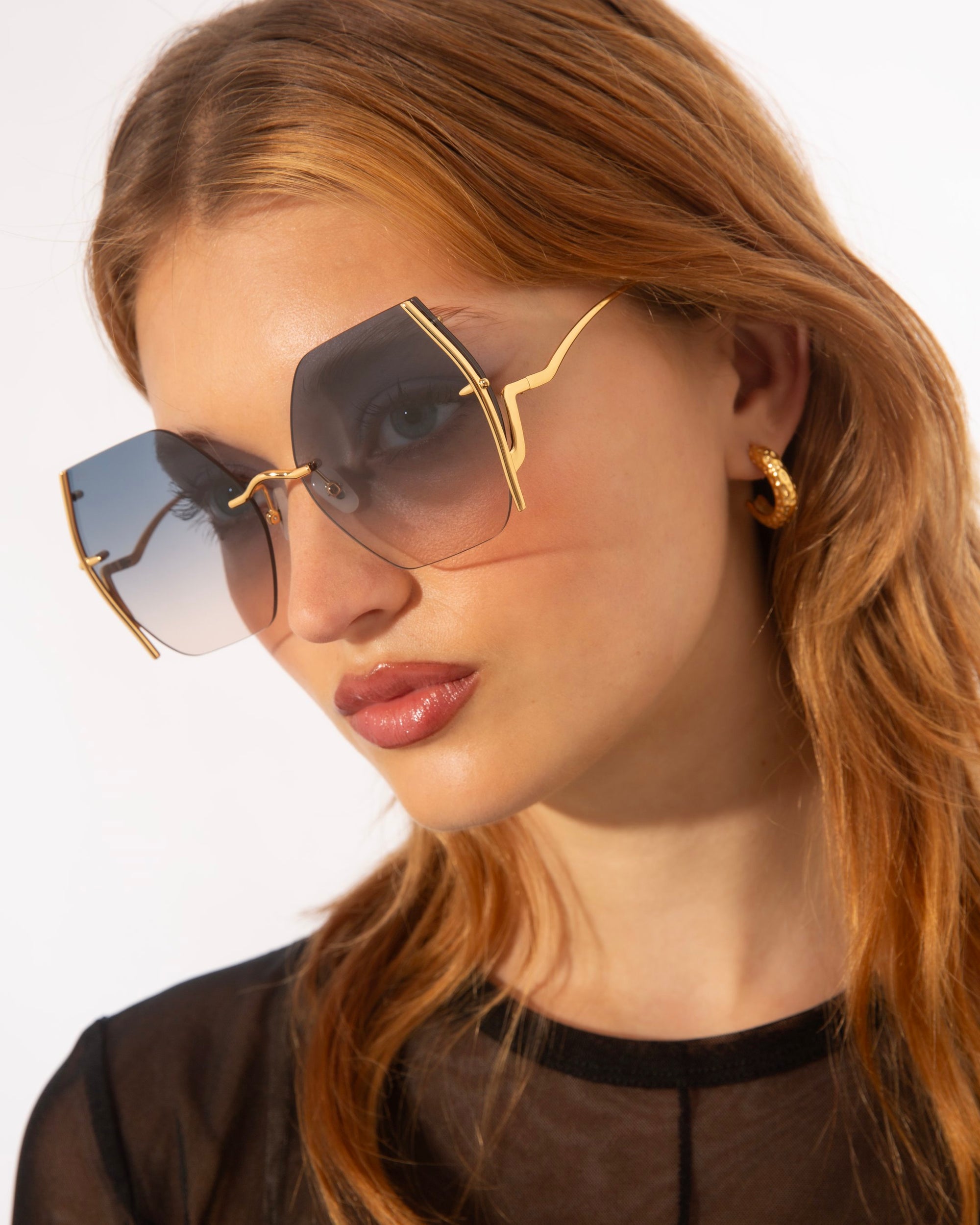 A woman with long, light brown hair wears oversized, square-shaped For Art's Sake® Generation sunglasses with a gold frame and jadestone nosepads for extra comfort. She has gold hoop earrings and is dressed in a black top. The For Art's Sake® Generation sunglasses offer UV protection, ensuring style and safety. The background is plain white.