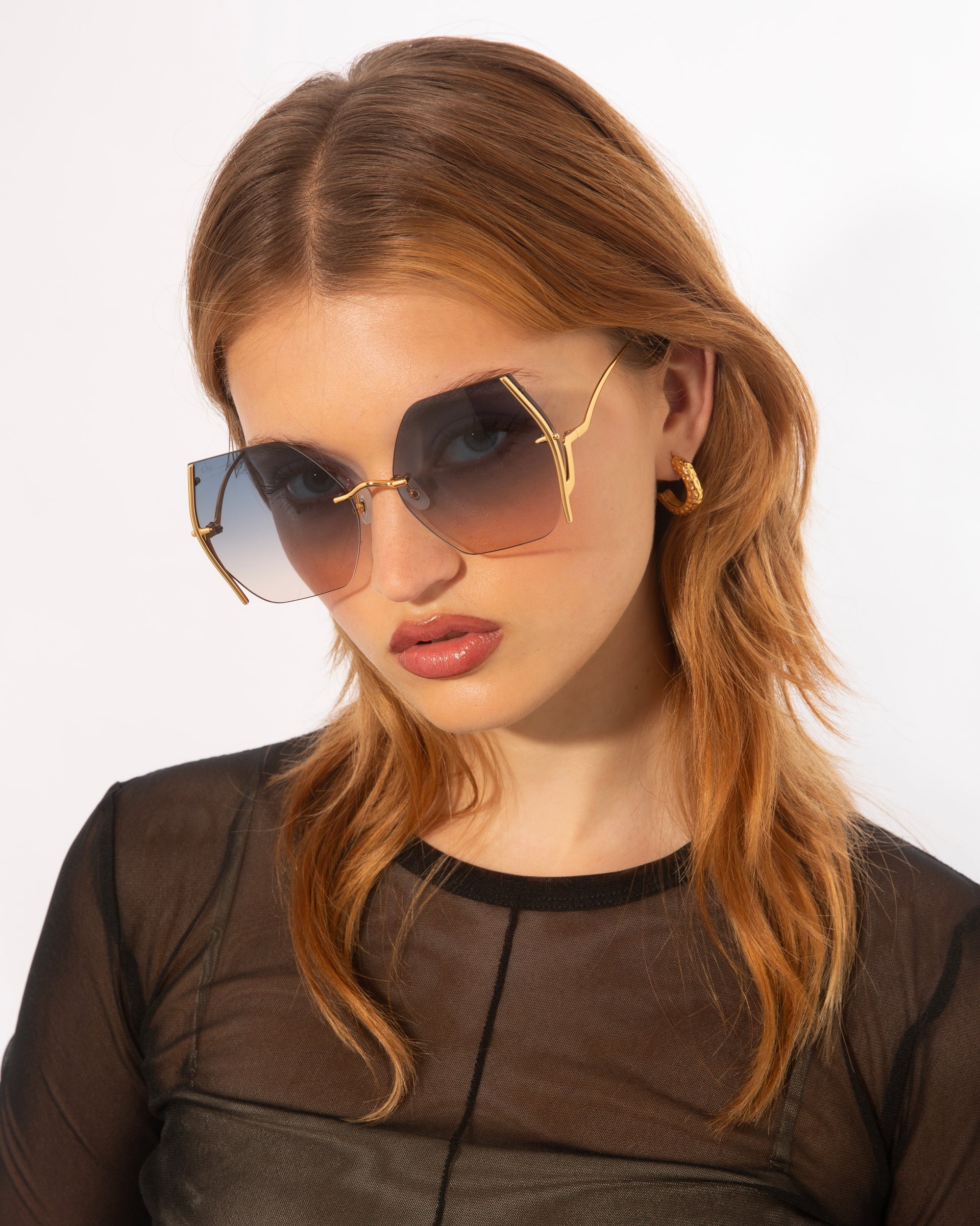 A woman with straight, auburn hair wears oversized, geometric sunglasses named Generation by For Art&#39;s Sake®, featuring gradient lenses and gold-plated stainless steel frames with jadestone nosepads for added comfort and UV protection. She has gold hoop earrings and is dressed in a black, sheer long-sleeve top. The background is plain white.