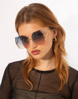 A woman with straight, auburn hair wears oversized, geometric sunglasses named Generation by For Art's Sake®, featuring gradient lenses and gold-plated stainless steel frames with jadestone nosepads for added comfort and UV protection. She has gold hoop earrings and is dressed in a black, sheer long-sleeve top. The background is plain white.