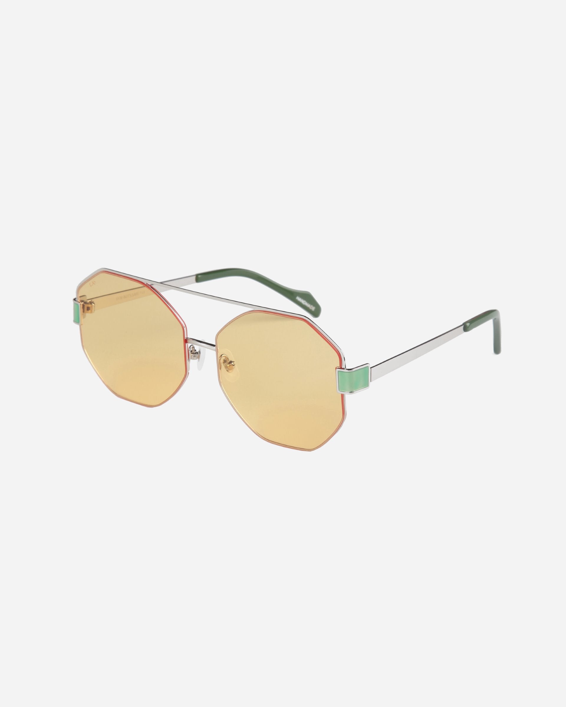 A pair of stylish For Art&#39;s Sake® Mania sunglasses with hexagonal, yellow-tinted nylon lenses, thin stainless steel frames, and green temple tips. The sunglasses have a retro vibe with a modern twist, featuring adjustable nosepads for comfort and a minimalistic design.