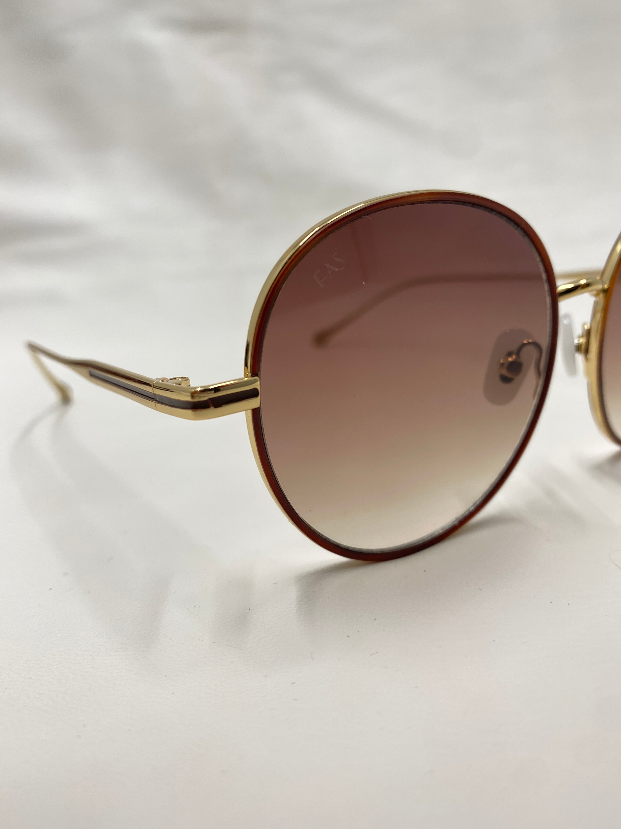 Close-up of the For Art's Sake® Lemon sunglasses featuring round, brown gradient lenses and thin, 18-karat gold frames. The background is a plain white surface.