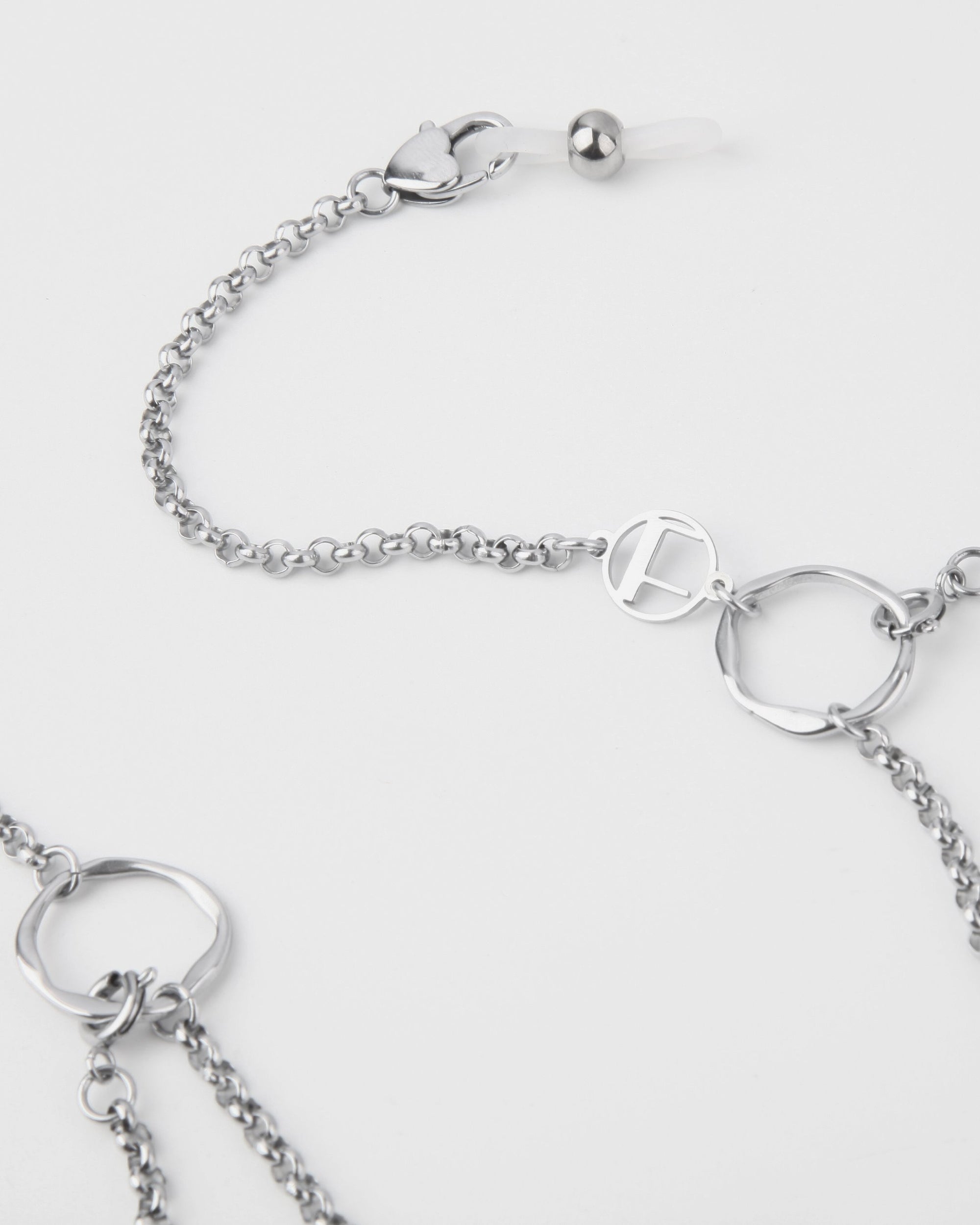 A close-up image of a For Art&#39;s Sake® Florentina Glasses Chain featuring various circular links, crafted from palladium-plated stainless steel. The glasses chain includes a decorative pendant with a stylized letter &quot;F&quot; and a clasp adorned with a small heart-shaped charm. The background is a clean white.